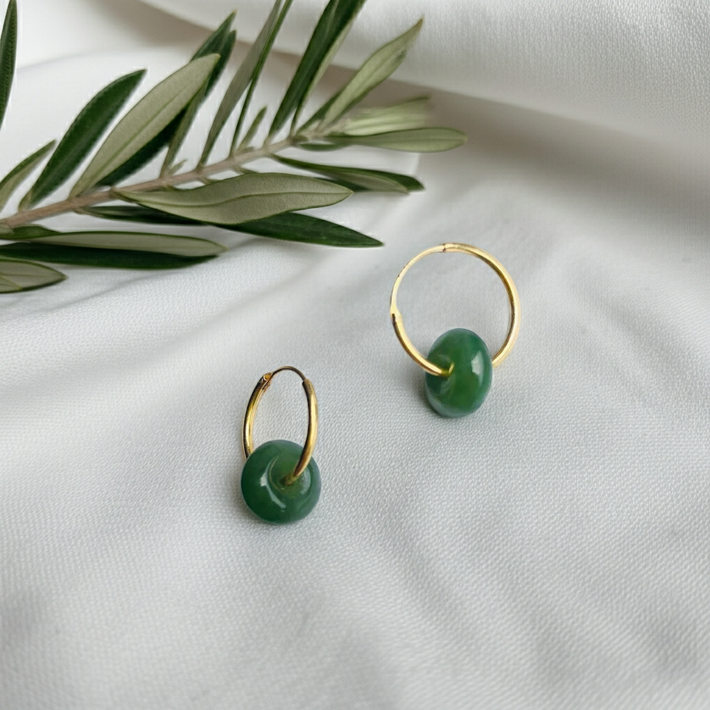 a pair of jade earrings with gold hoop on top of white fabric and olive leaves decoration