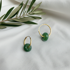 a pair of jade earrings with gold hoop on top of white fabric and olive leaves decoration