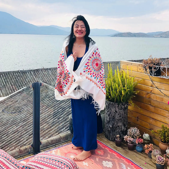 Robin - owner of SpringCity Dream standing on a balcony with a scenic view of water and mountains, holding a colorful shawl, wearing silver jewelry and has a big smile on her face,