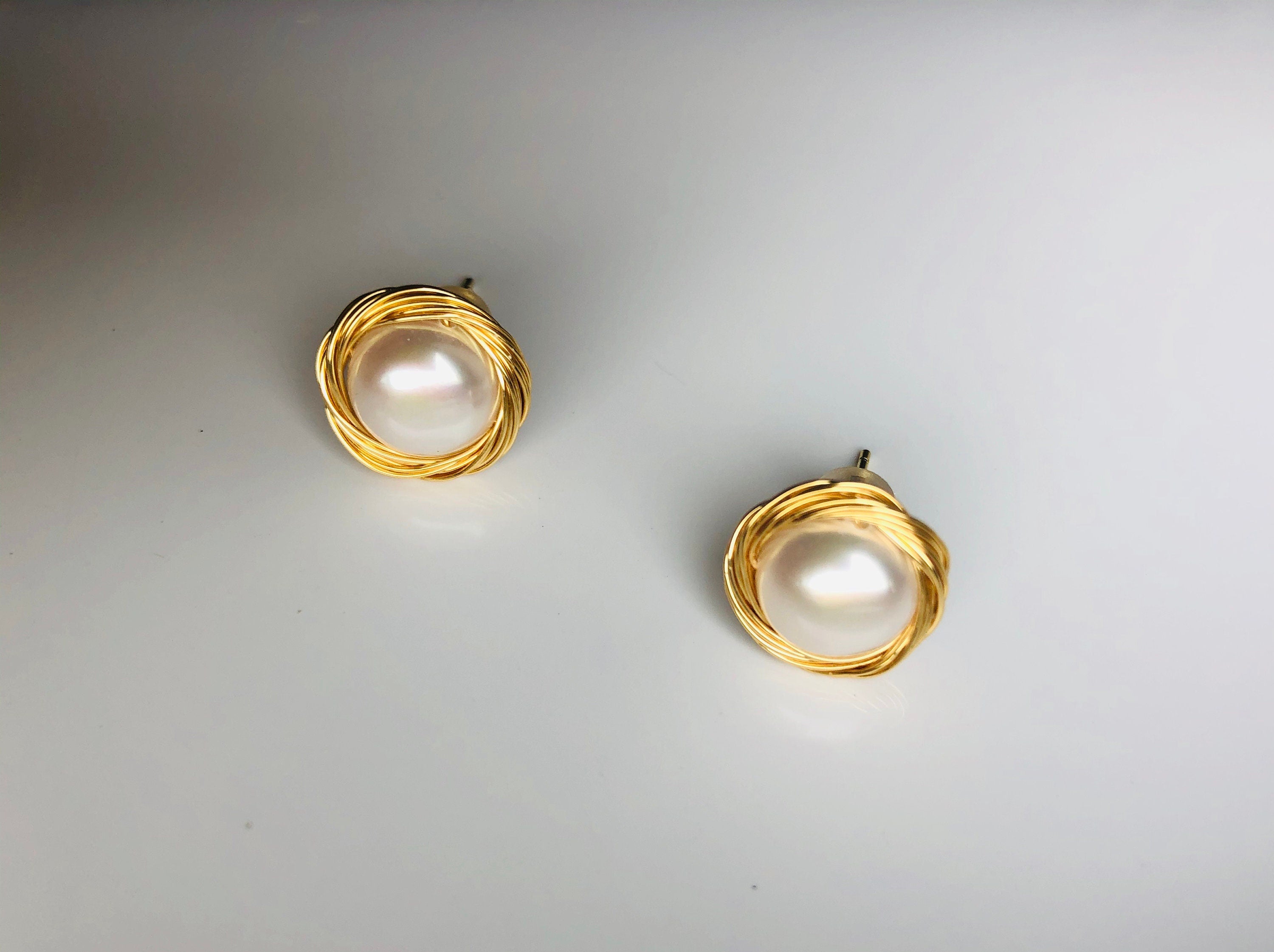 A pair of gold-filled wired pearl stud earrings placed on a white surface.