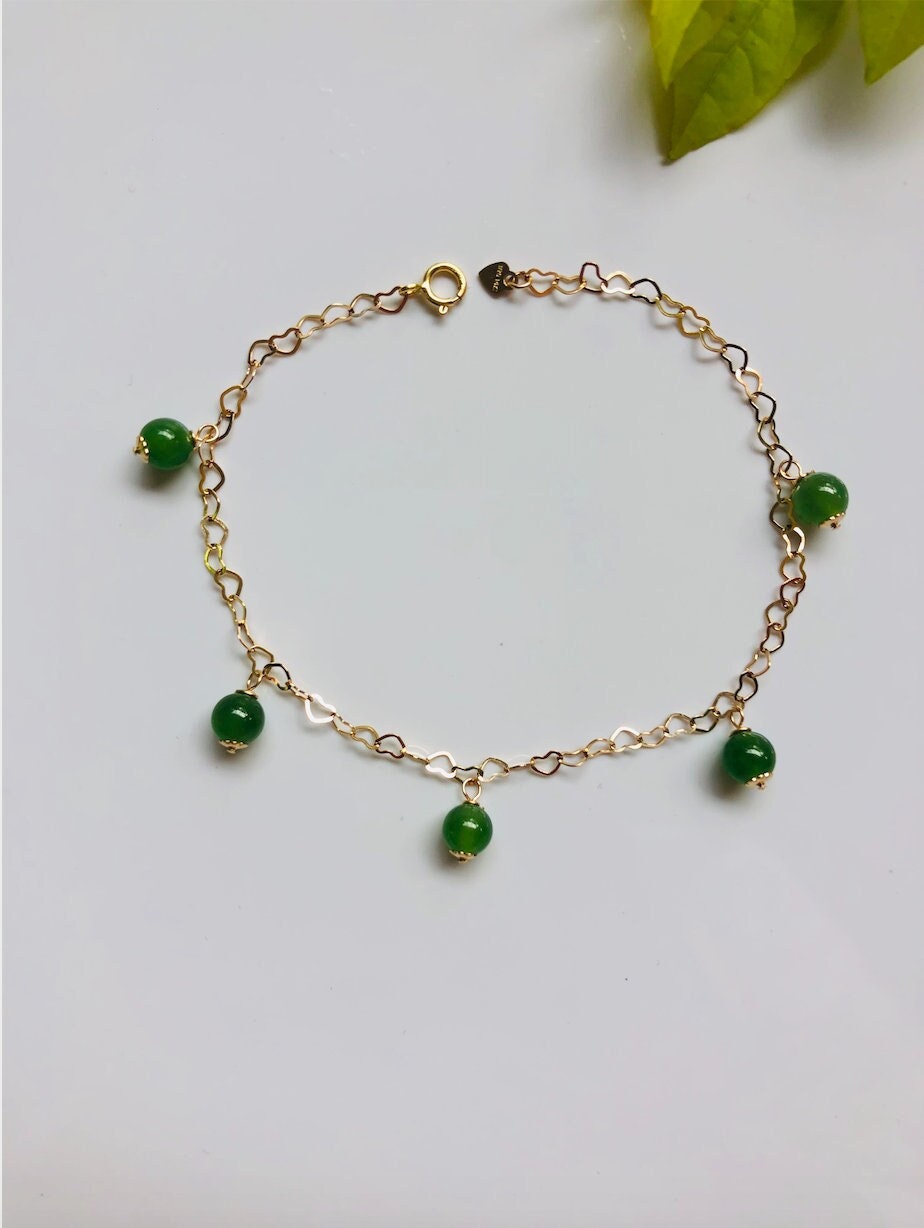 An adjustable gold-colored bracelet featuring heart-shaped charms and green jade beads laid out on a flat surface.