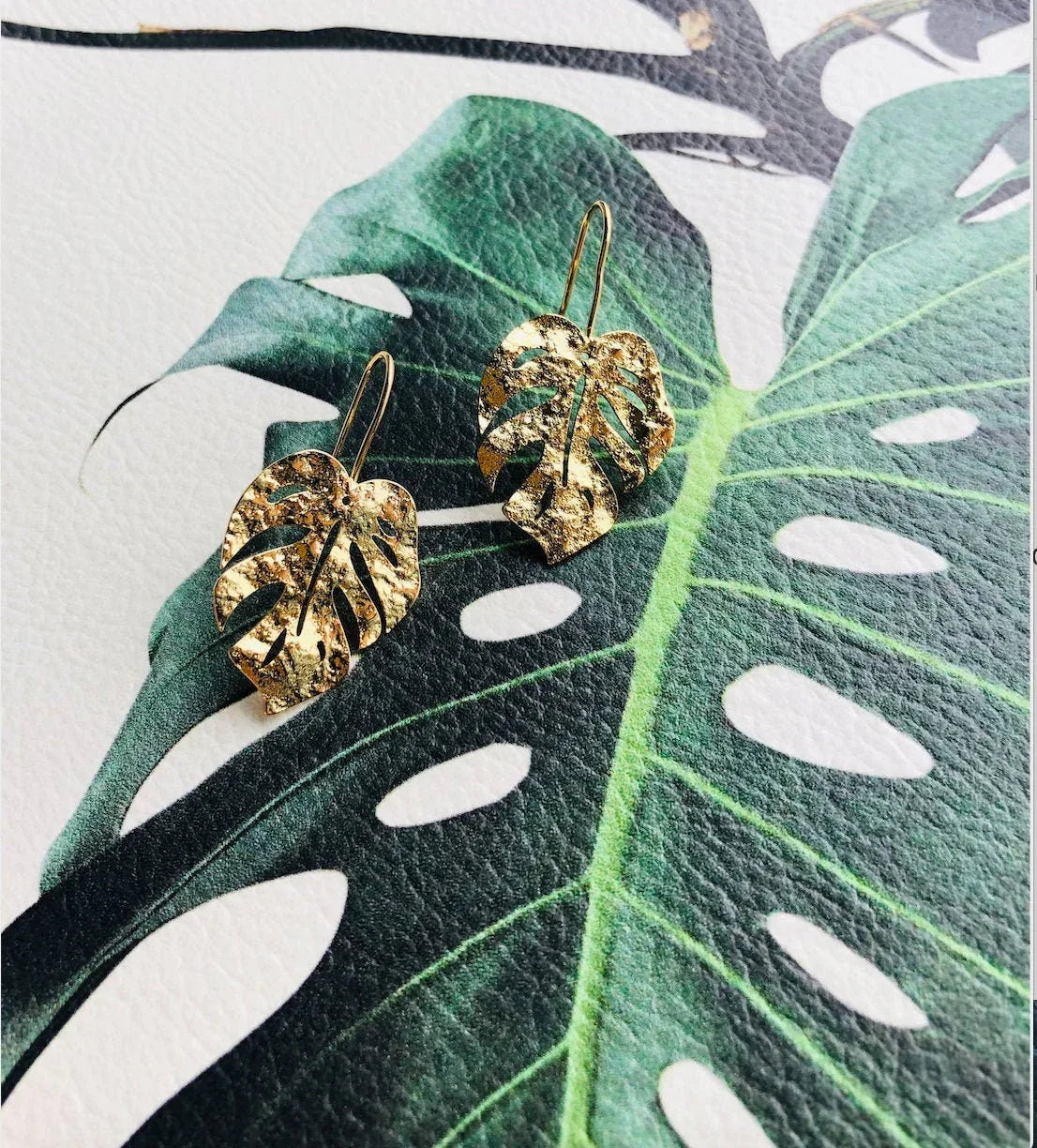 A pair of gold-plated Monstera leaf earrings displayed on a green leaf background, highlighting their tropical design.