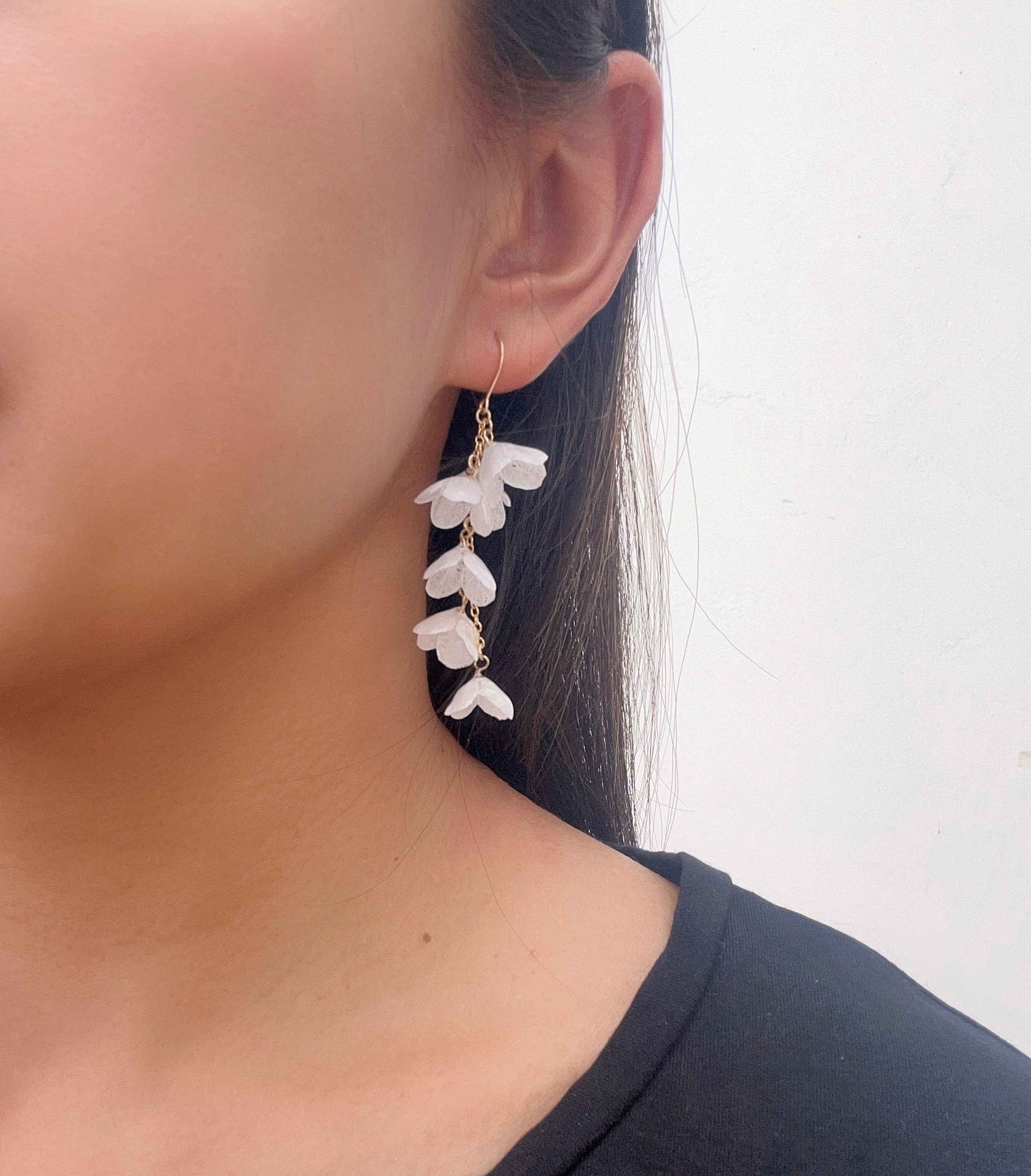 A person wearing a pair of asymmetrical gold hanging earrings with white preserved flower petals attached, in an irregular pattern.