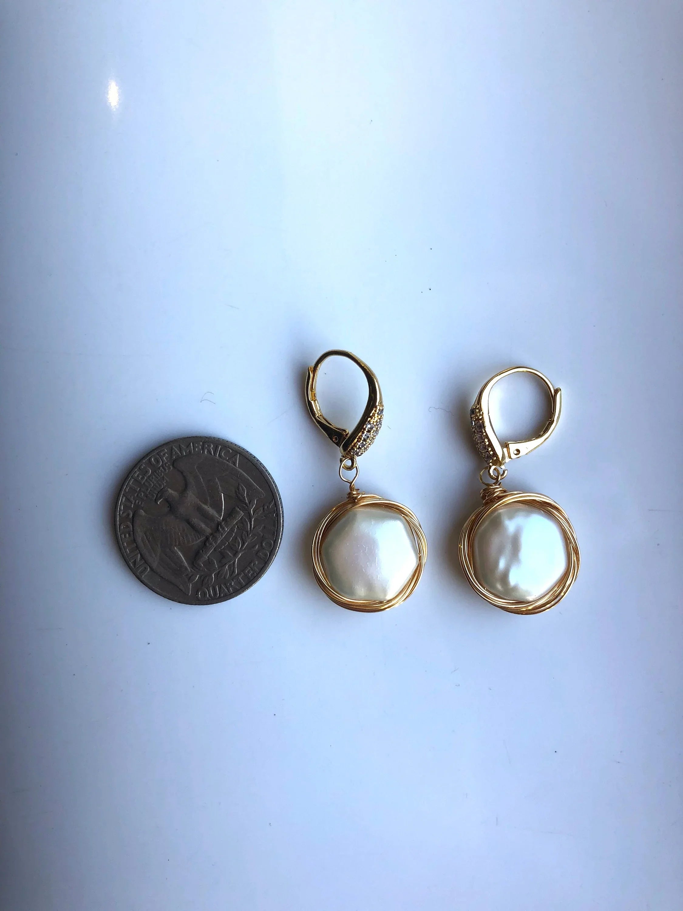 Pair of pearl earrings with gold accents ext to coin for size and proportion reference on a white background