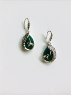 A pair of teardrop-shaped dark green jade earrings with silver peacock designs and red/ruby stone eyes, attached to antique silver hooks.