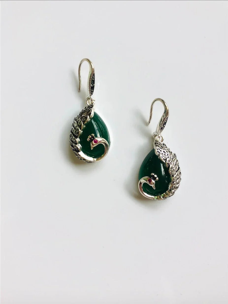 A pair of teardrop-shaped dark green jade earrings with silver peacock designs and red/ruby stone eyes, attached to antique silver hooks.