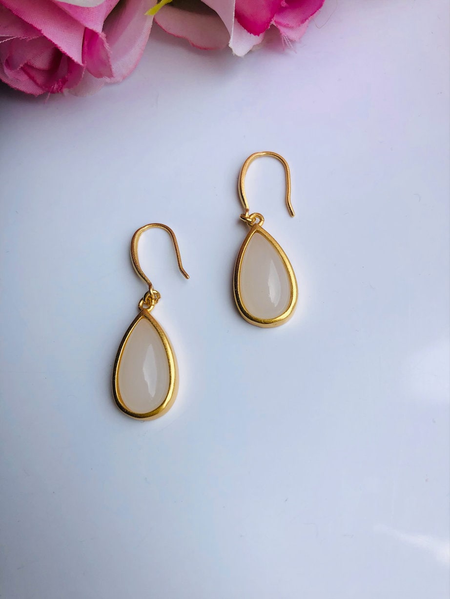 A pair of teardrop-shaped white jade earrings with gold accents, displayed against a floral background.